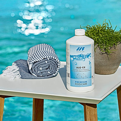 Flasche ALG-EX steht auf Tisch; daneben Handtuch, Pflanze; Hintergrund: verschwommener Pool.