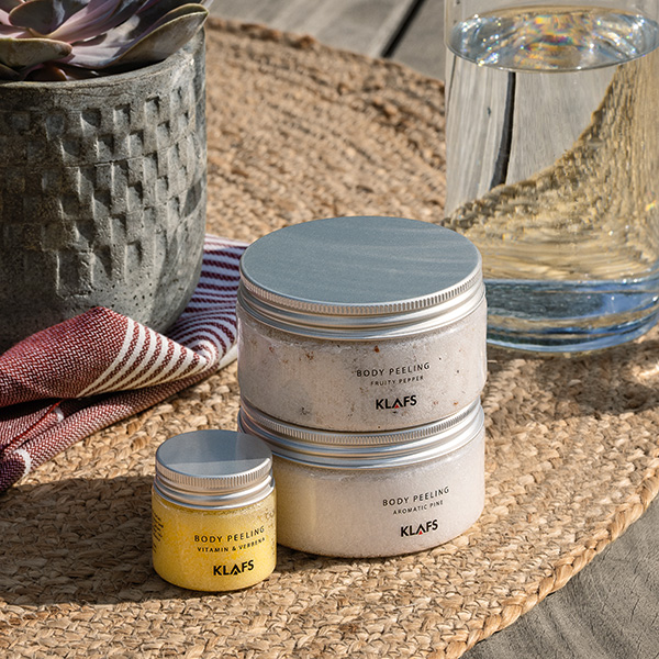 Drei Dosen Body-Peeling stehen auf einem geflochtenen Untersetzer neben Glas und Pflanze draußen.