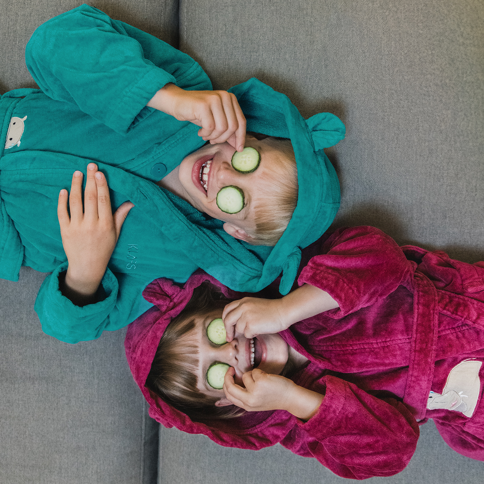 Kinder liegen lachend auf Sofa, tragen Bademäntel und halten Gurkenscheiben vor den Augen.
