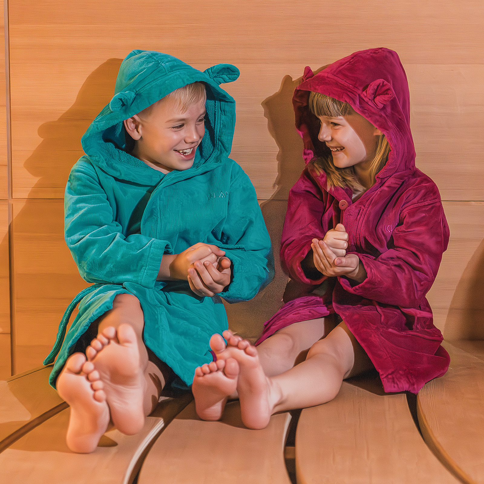 Kinder sitzen lachend in bunten Bademänteln auf einer Saunabank aus Holz.