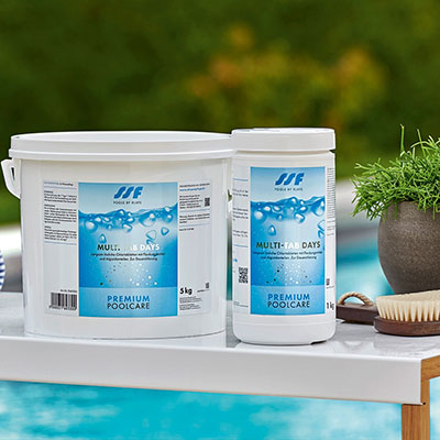 Zwei Eimer Poolpflegeprodukte stehen auf einem Tisch am Pool, umgeben von Pflanzen.